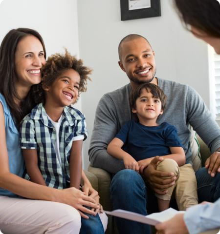 Young couple sitting in chairs with their two sons as a woman holding a piece of paper explains something to them.