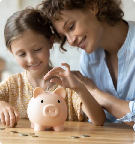 woman sitting with a little girl who is putting coins in a pink piggy bank.