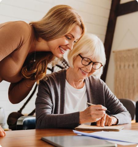 young woman leaning over elderly woman as she writes something.